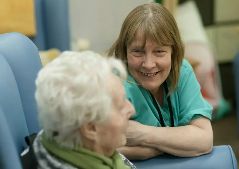 A smiling caregiver in scrubs talks with a seated older woman